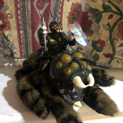 A fuzzy, brown black and green spiderlike mount with orange eyes, with a spiderlike humanoid rider sitting atop, holding a large sickle and shield, with a sword at his side.