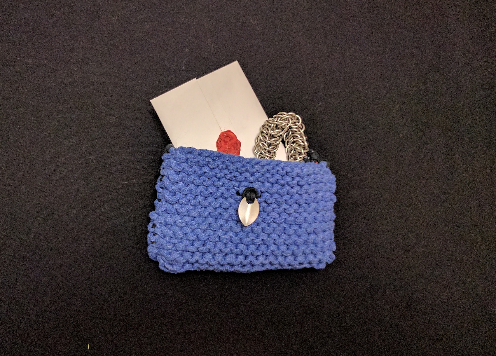 A blue knitted envelope on a black surface, with a wax sealed envelope and chainmaille mobius strip inside.