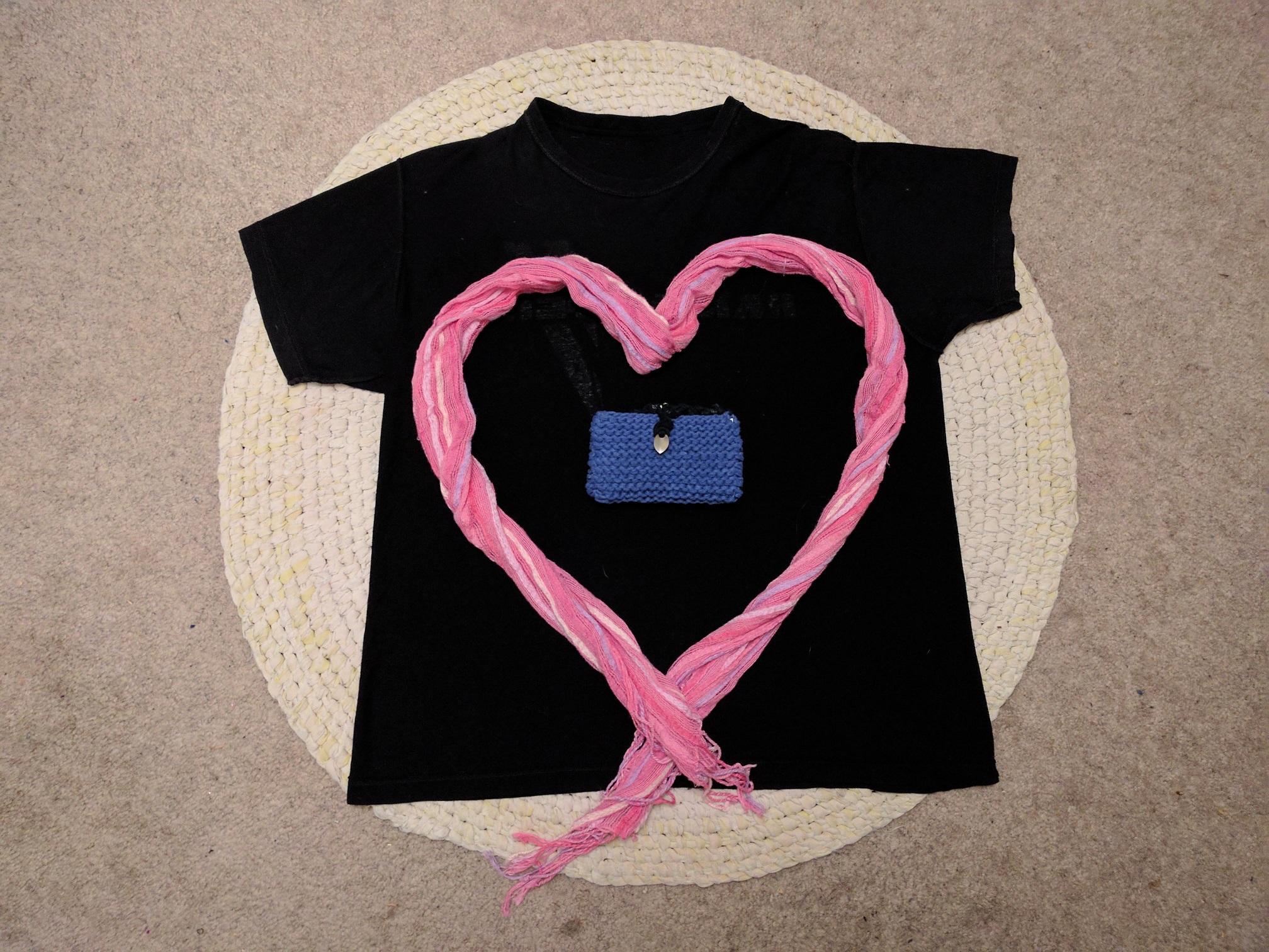 A tshirt on a knitted rug, with a pink heart drawn in the center with a scarf, and the blue envelope in the middle.