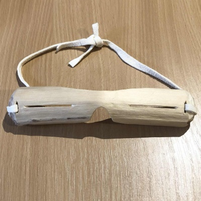 An pair of willow wood snow googles, with wide, thin slits to see through, with white leather straps, laying on a wooden table.