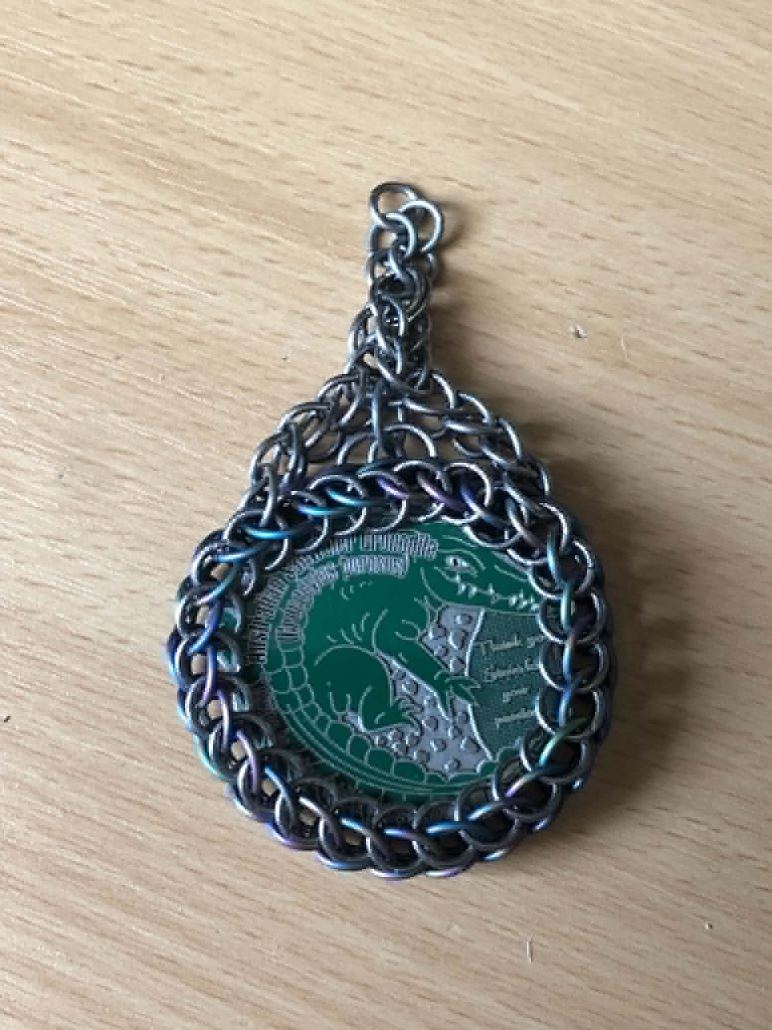 A chainmaille wrapped coin on a wooden table, using silver rings and rainbow anodized rings, with a chain attached.
