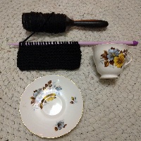 The beginnings of knitting a cup and saucer cozy, with the saucer and cup layed out beside. The knitted portion is the width of the saucer, but only about a third of the way tall