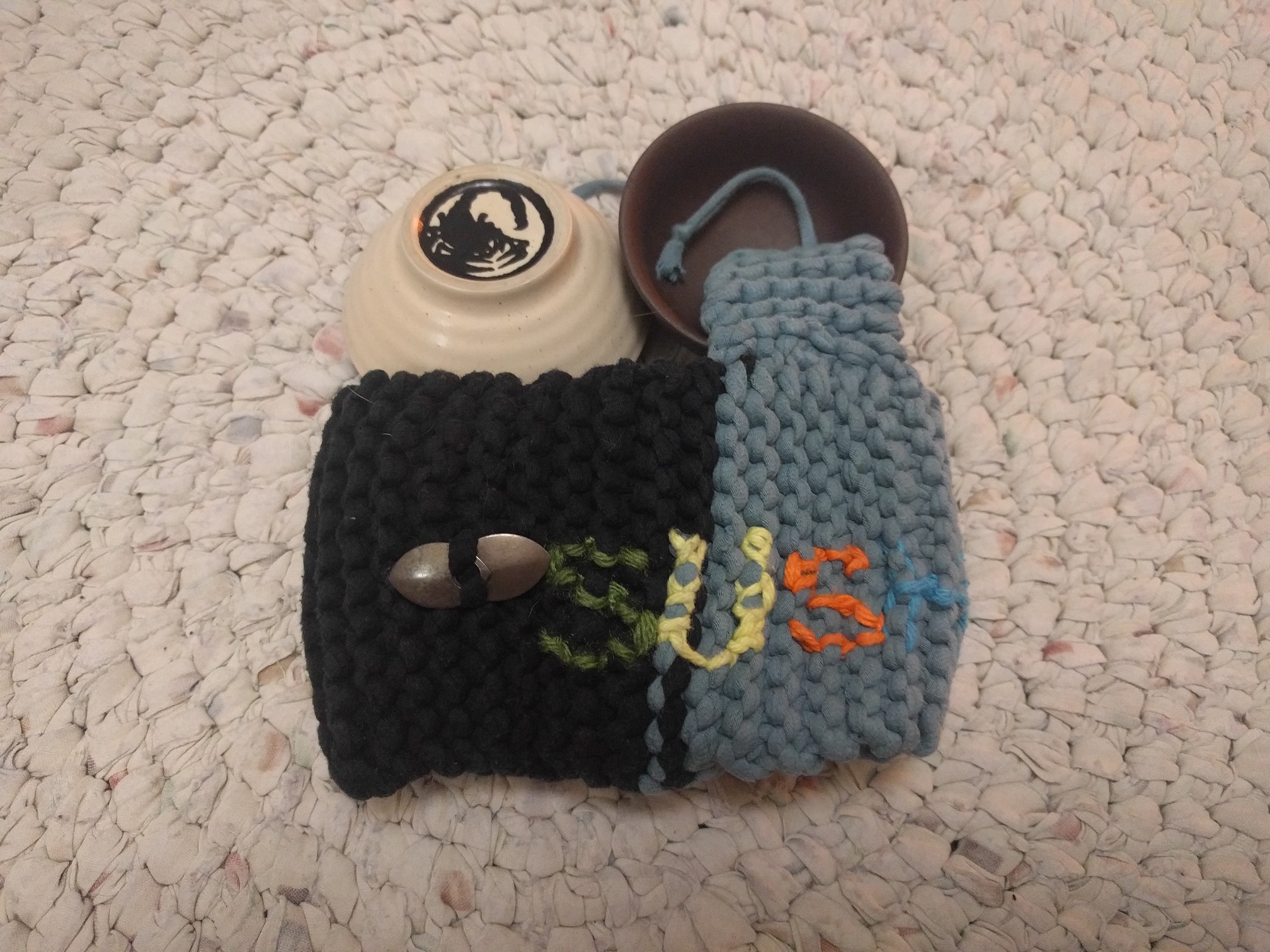 A teacup cozy in black and blue, with the first few letters S U S H on the front, and the two cups removed from it, all of it laying on a crocheted rug.