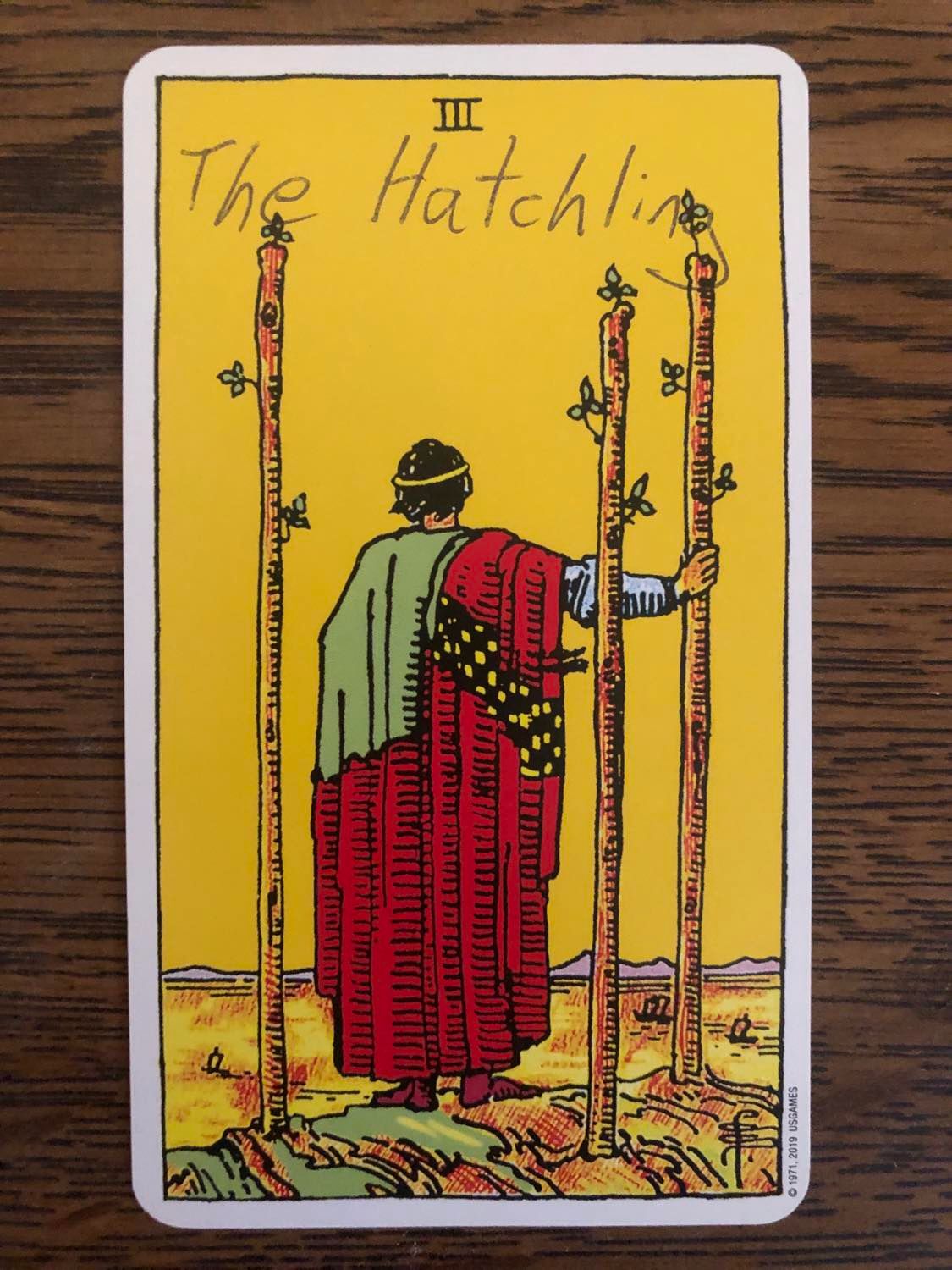 The three of wands card with the hatchling written on it, laying on a wooden desk.