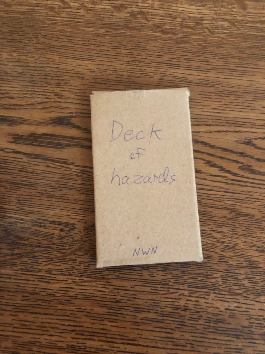 The Deck of hazards box, raw cardboard with the name written on the front, laying on a wooden table.