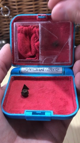 A still image from the video of a dice rolling through the miniature clear dice tower, with the d20 visible inside, part way through tumbling down the tower. The inside is lined with red velvety fabric, with a small dice bag tucked into the upper left beside the tower, a sparkly blue spell scroll for Haste is inside the hinge space, and a d12 and d4 is laying inside the lower tray where the dice land.