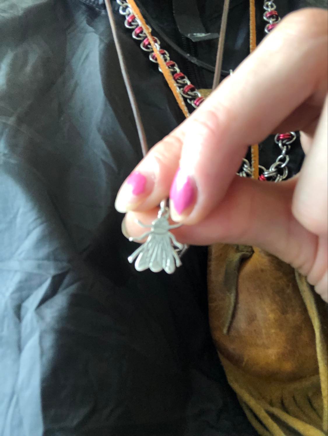 Kabutroid holding up the spiritual housefly pendant around her neck, with the Rosary, Medicine bag, and Crow pendant behind, and wearing a black windbreaker for the day.
