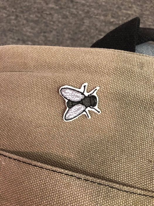 A grey housefly patch with a white edging, sewn onto a grey canvas computer bag, placed on the back near the top, generally facing towards the handle of the bag.