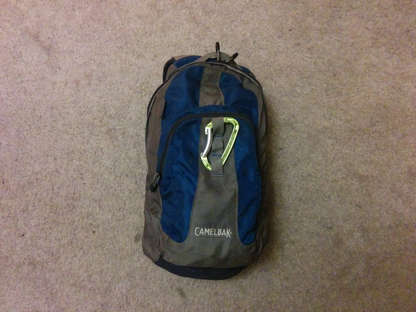 A blue and grey backpack laying on an offwhite carpet, with a green mountaineering caribiner clipped to the outside.