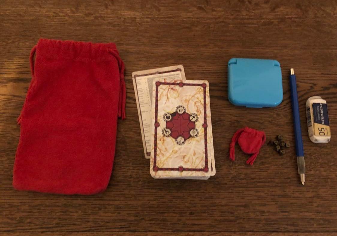 The red harrow bag with the deck to the right of it. The closed dice tower case is to the right of it, with the tiny dice bag and dice beneath. To the right of those is a mechanical pencil and an eraser to the right of that.