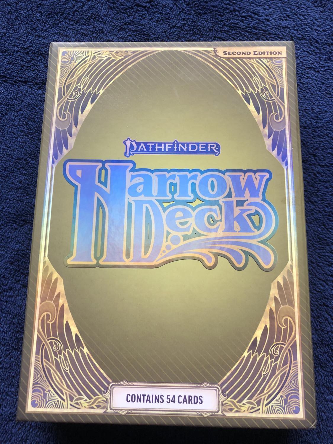 The card box, in shiny gold with purple lettering. The box like the cards in 2e have a wing-like surround to the images, and is much larger than the cards themselves. The cards inside are held in the center with cardboard borders, and the instruction manual is the size of the box itself, which is in the next pic.