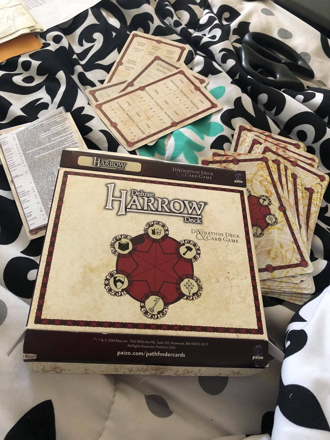 The original box, which is two cards wide for some reason. It is layong on top of the deck of many things card, the actual divination instruction cards, and the harrow cards themselves. The box says 'Deluxe Harrow Deck Divination Deck & Card Game' in grey text on a light brown background, with the card's logo image underneath. The logo itself is a dark brown six pointed star inside of a circle, with each corner being the symbols for each of a character attributes, that being a crown for charisma, a book for intelligence, a hammer for strength, a shield for constitution, a star for wisdom, and a key for dexterity.