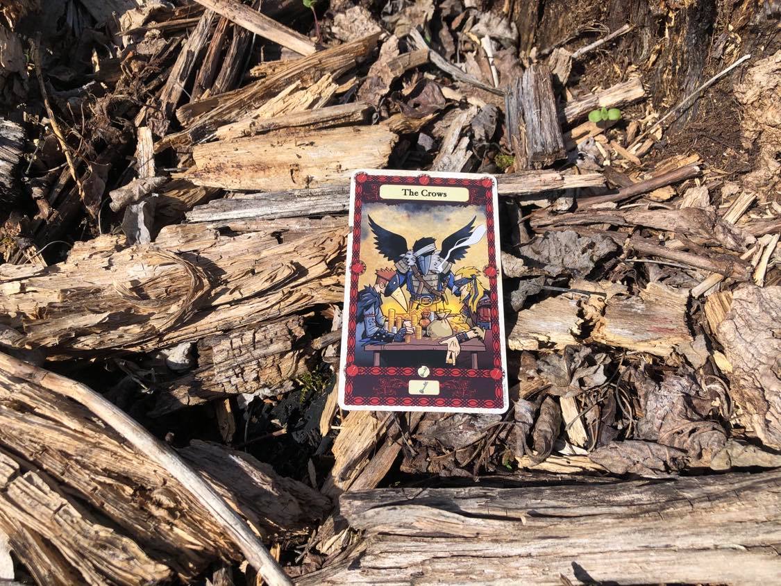 Two men and a humanoid crow, the card laying on the forest debris.