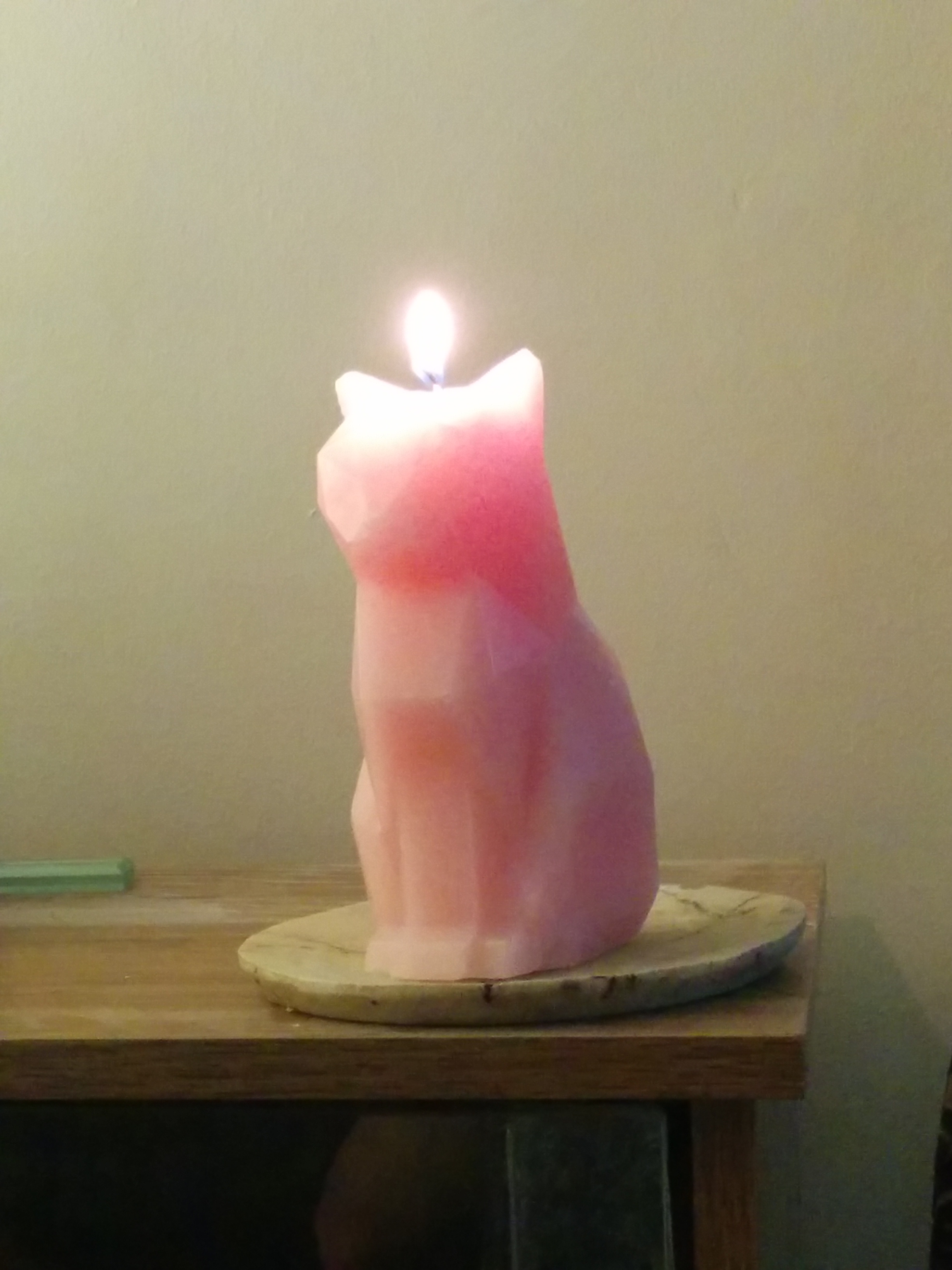 A pink cat-shaped candle burning on a stand, on the corner of a livingroom cabinet.