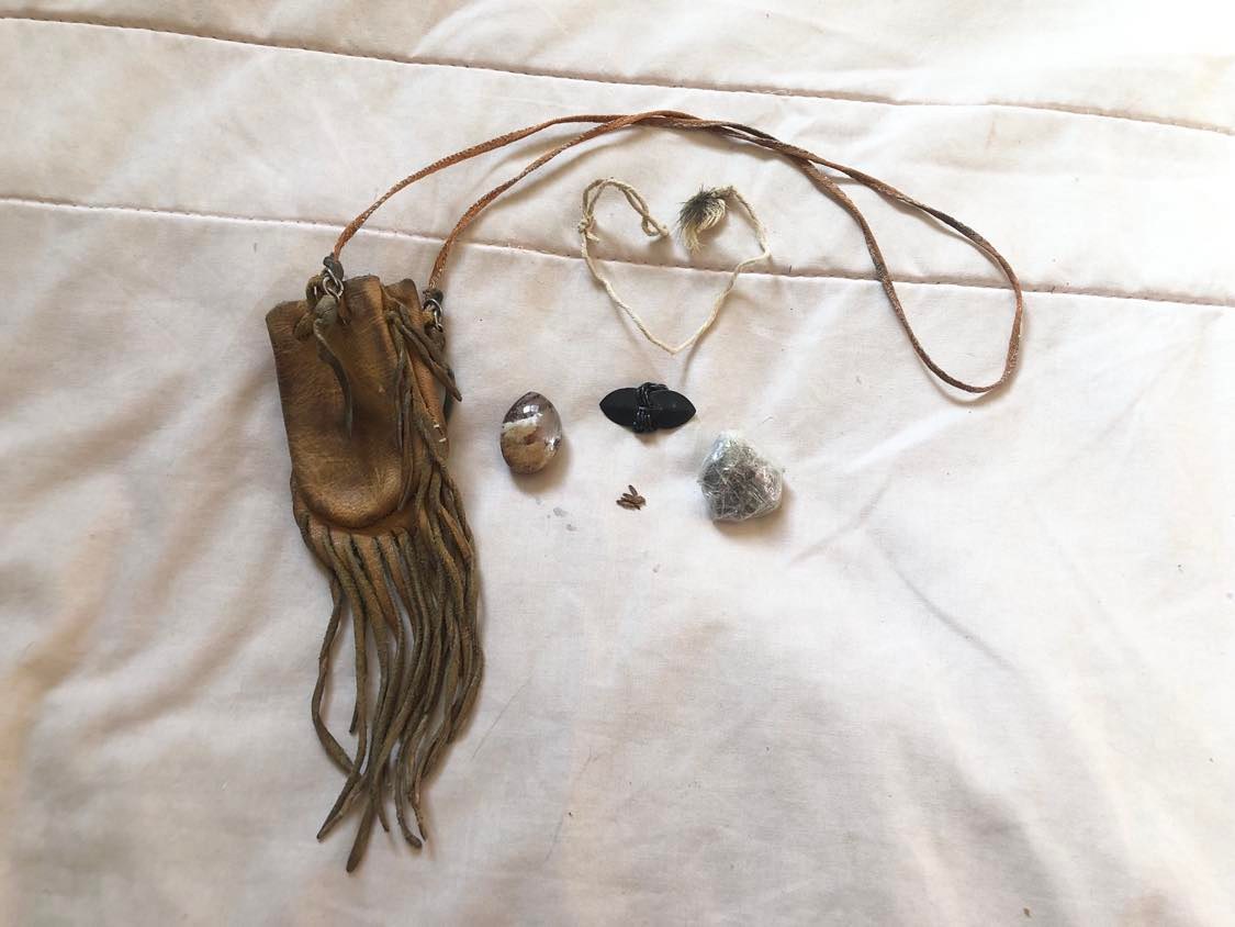 The pouch with string toy, Pagan crow, the Jack stone, a sweatlodge rock, and a little pile of tobacco are sitting on an offwhite blanket. The string and feather toy is in the shape of a heart.
