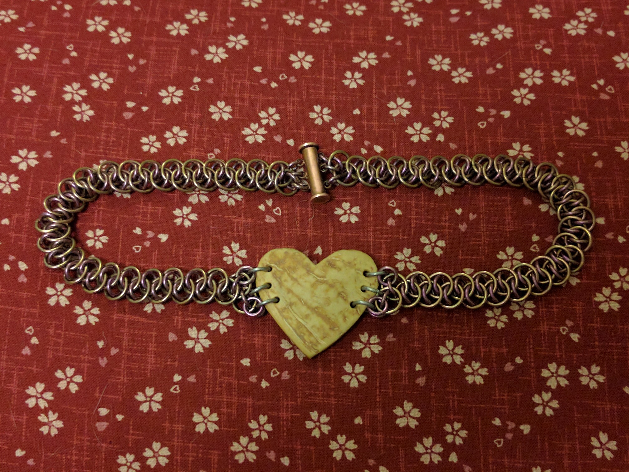 The heart pendant, attached to the chainmaille collar, sitting on a sakura pattern burgundy bed.