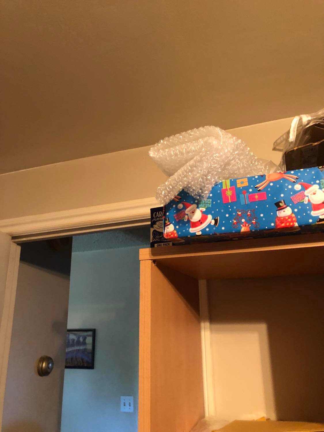 A blue card box, hidden beside a wrapped box and some bubblewrap on top of a shelving unit, though precariously placed, almost as though deliberately.