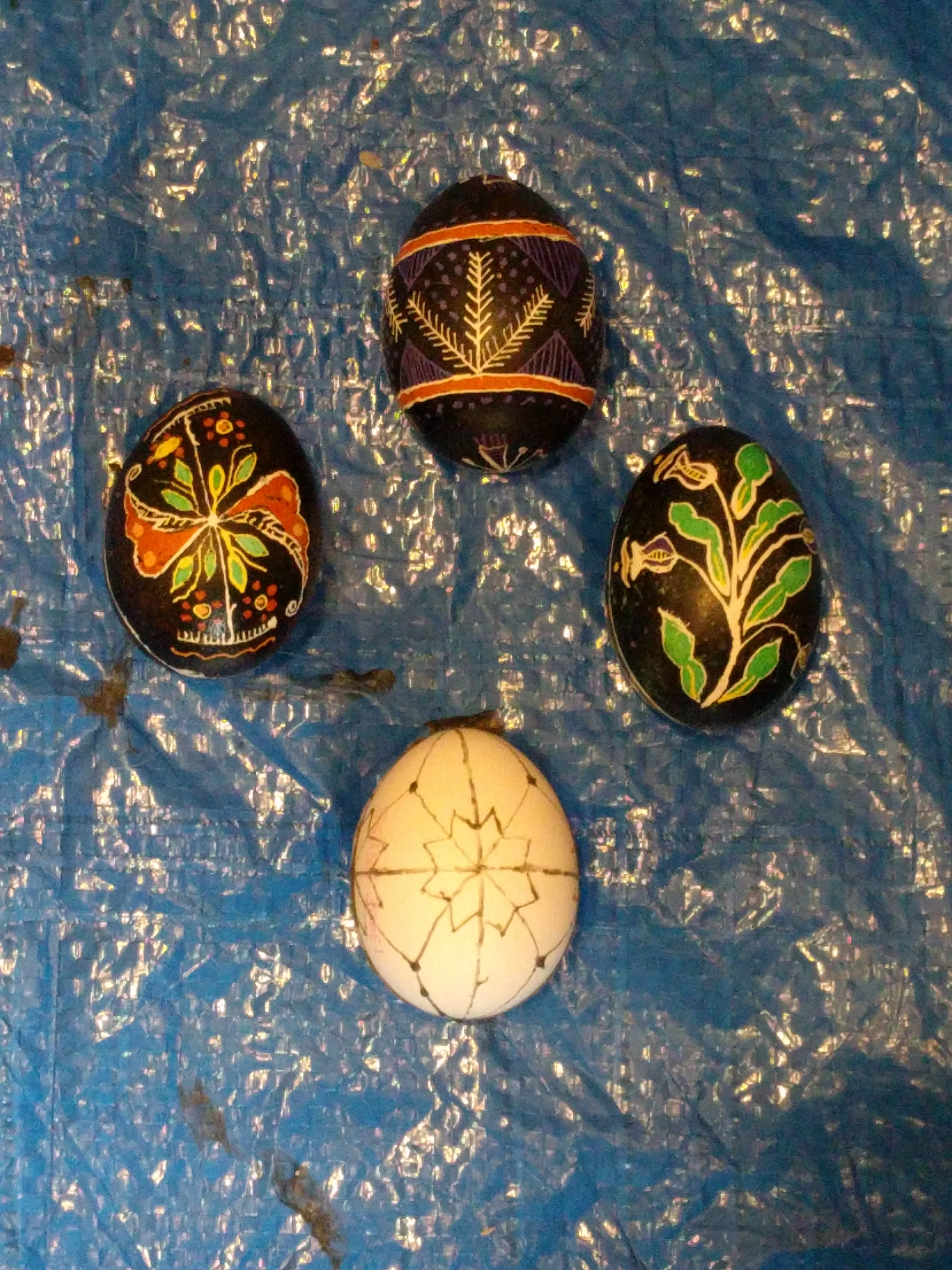 Four eggs on a blue tarp, flowery or with wheat sheaves, and one that isn't yet painted, but has some wax applied.