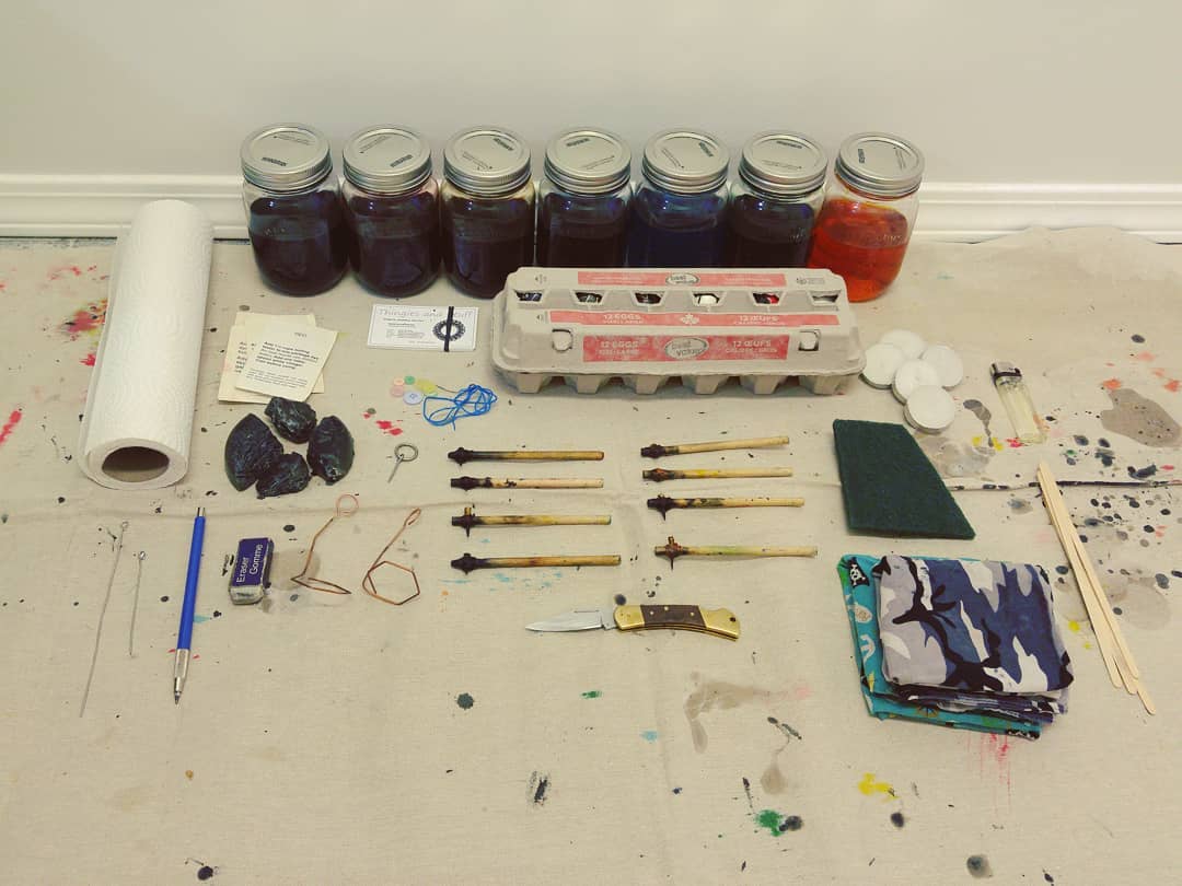 Our egg-painting kit, with eight syluses, seven colours prepared, paper towel, the wax, a carton of hollowed out eggs, and other supplies arranged on the canvas worktop.