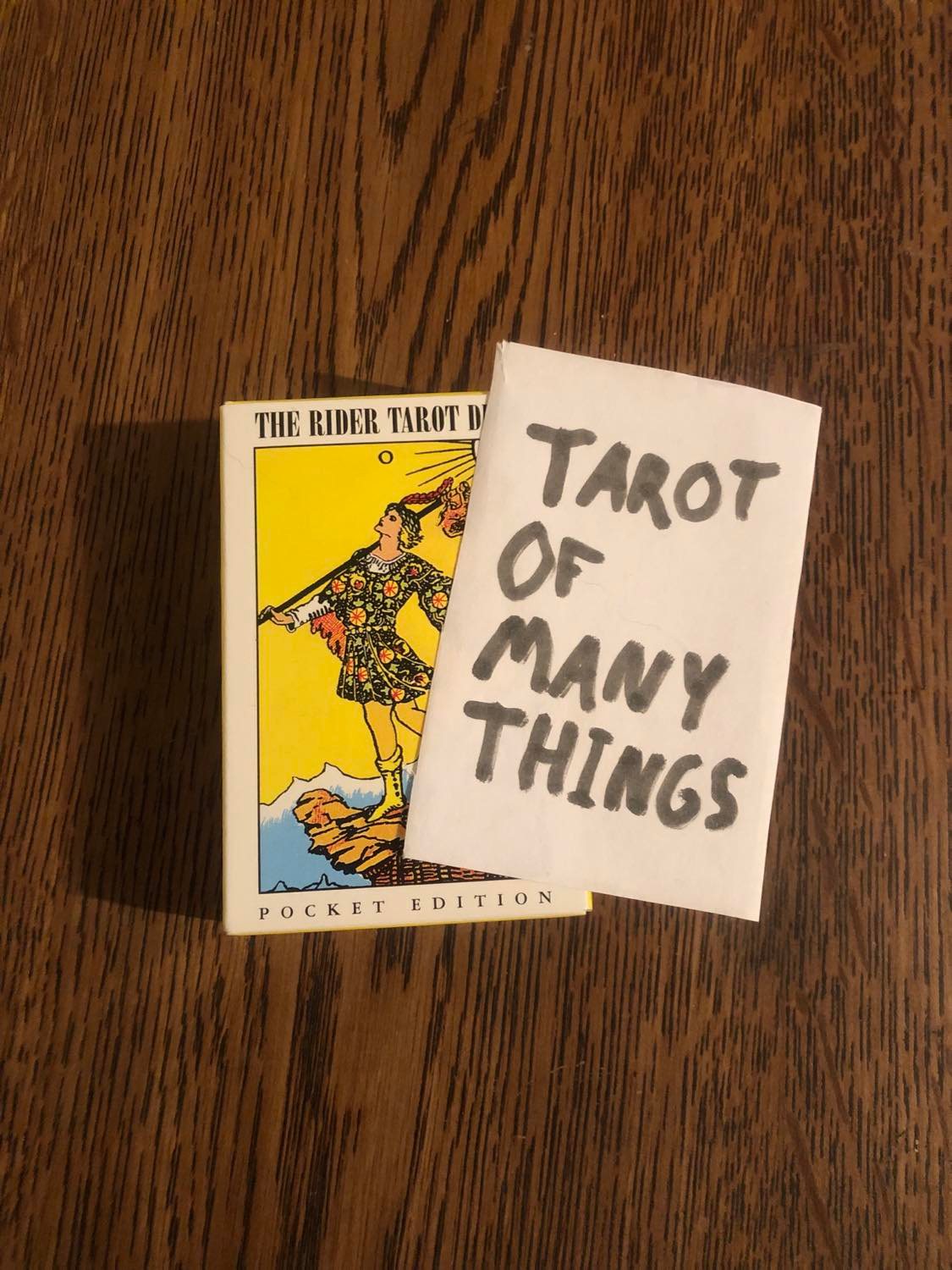 A scratchily markered paper saying Tarot of Many Things, on top of a rider waite tarot deck box, sitting on a wooden style table.
