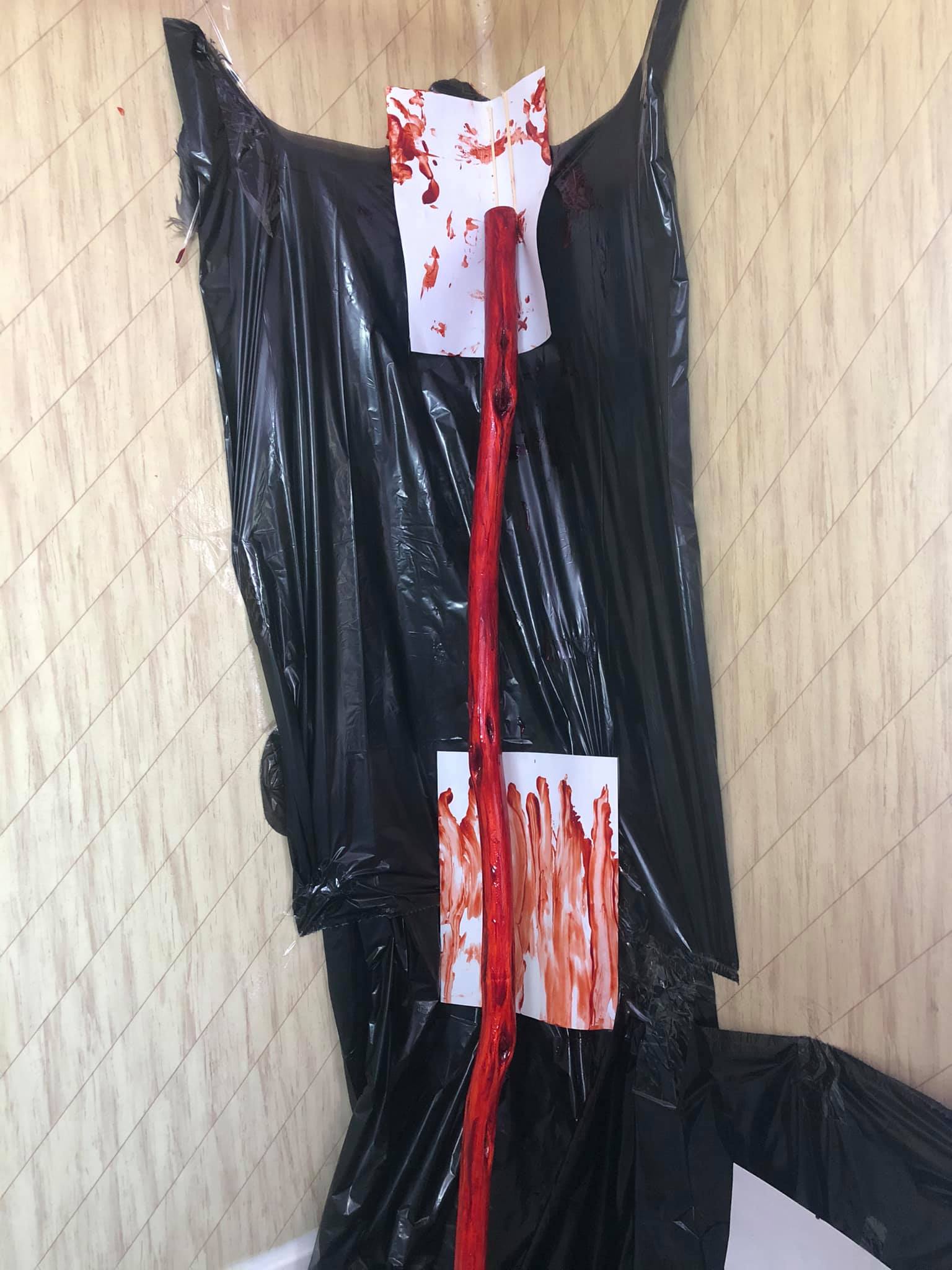 A red staff leaning up against a corner of a bathtub wall, black garbage bags painted to the wall to protect them. Bizarre, blood covered canvases are behind the staff.