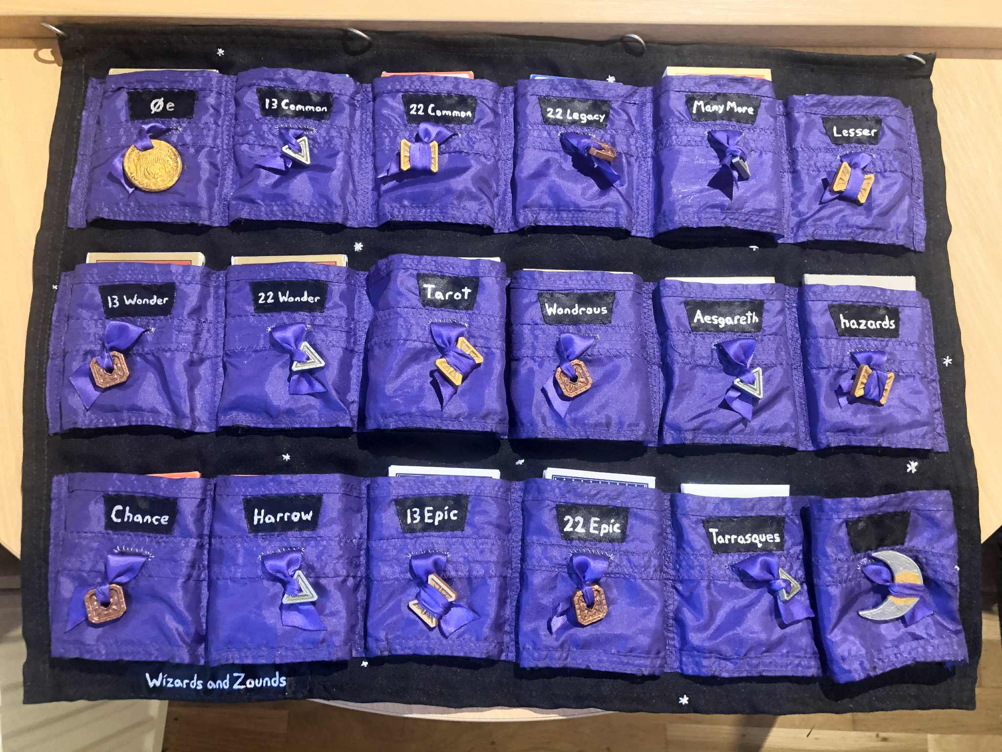 The cardsleeve, fully laid out on a table, each of the pockets filled with their respective decks, with the exception of the very last pocket as only seventeen decks have been discovered, three of them being homebrew. Silver sparkles dot the fabric, labelled as belonging to Wizards and Zounds on the bottom left.