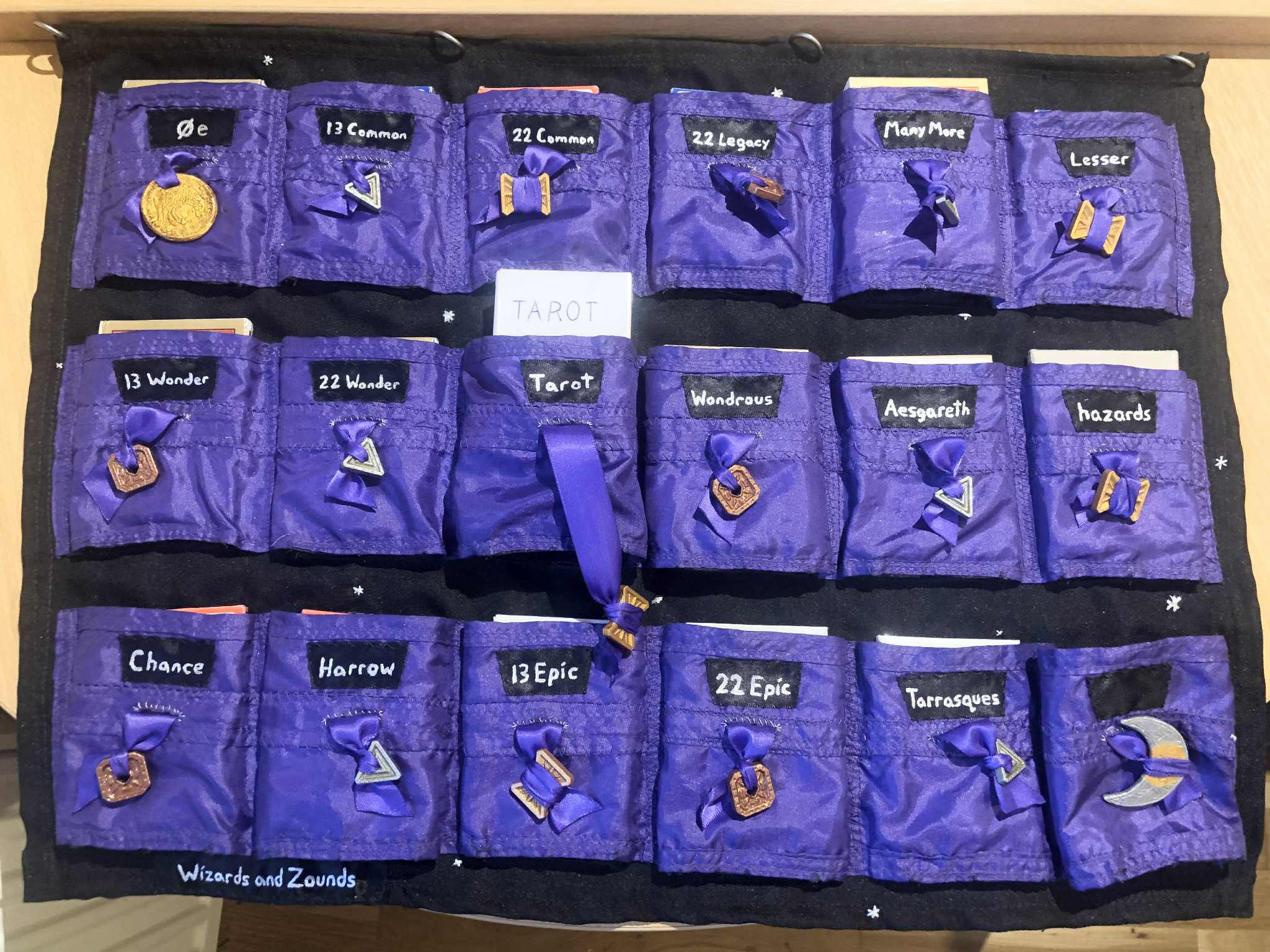 A fabric sheet with many pockets, eighteen total in three rows, though the last row has no label on the last pocket. Shimmery purple fabric on black, each with a drawstring tipped with a dungeons and dragons coin, and the pockets labelled things like zero e, 13 common, 22 wonder, wondrous, harrow, 22 epic, and more. All of the random draw type decks of many things from Dungeons and Dragons.