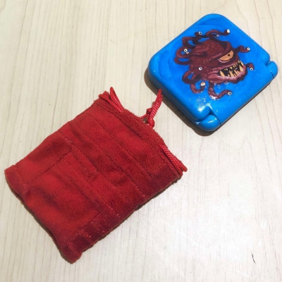 A red velvety pouch made from lots of thin red scraps of fabric, with the open side facing towards a small painted blue box that would fit perfectly inside.