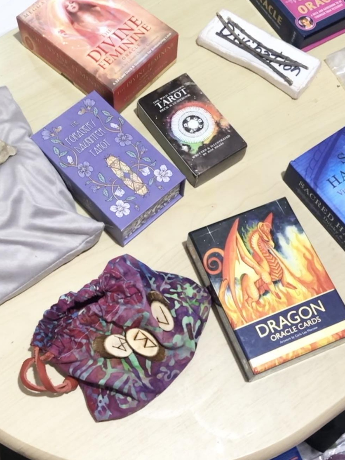 An assortment of tarot decks, oracle decks, and divination sets spread about on a table, some being the forager's daughter tarot, dragon oracle cards, a set of three divination sticks, and others.