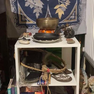 A white table tucked between a sewing desk and the wall radiator, containing a smudge pan, singing bowl, two stones saying peace and faith, a mjolnir hammer, a glass bear, a sweetgrass braid, sage, and books and angels can be seen beside the table.