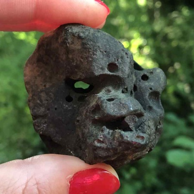 A dark grey chunky stone that strongly resembles a primape's face, with the hole through the stone being located exactly where the primape's right eye would be. A formation coming off the side of the face strongly resembles a turtle head as well, and it's being held up in front of a backdrop of a forest canopy.