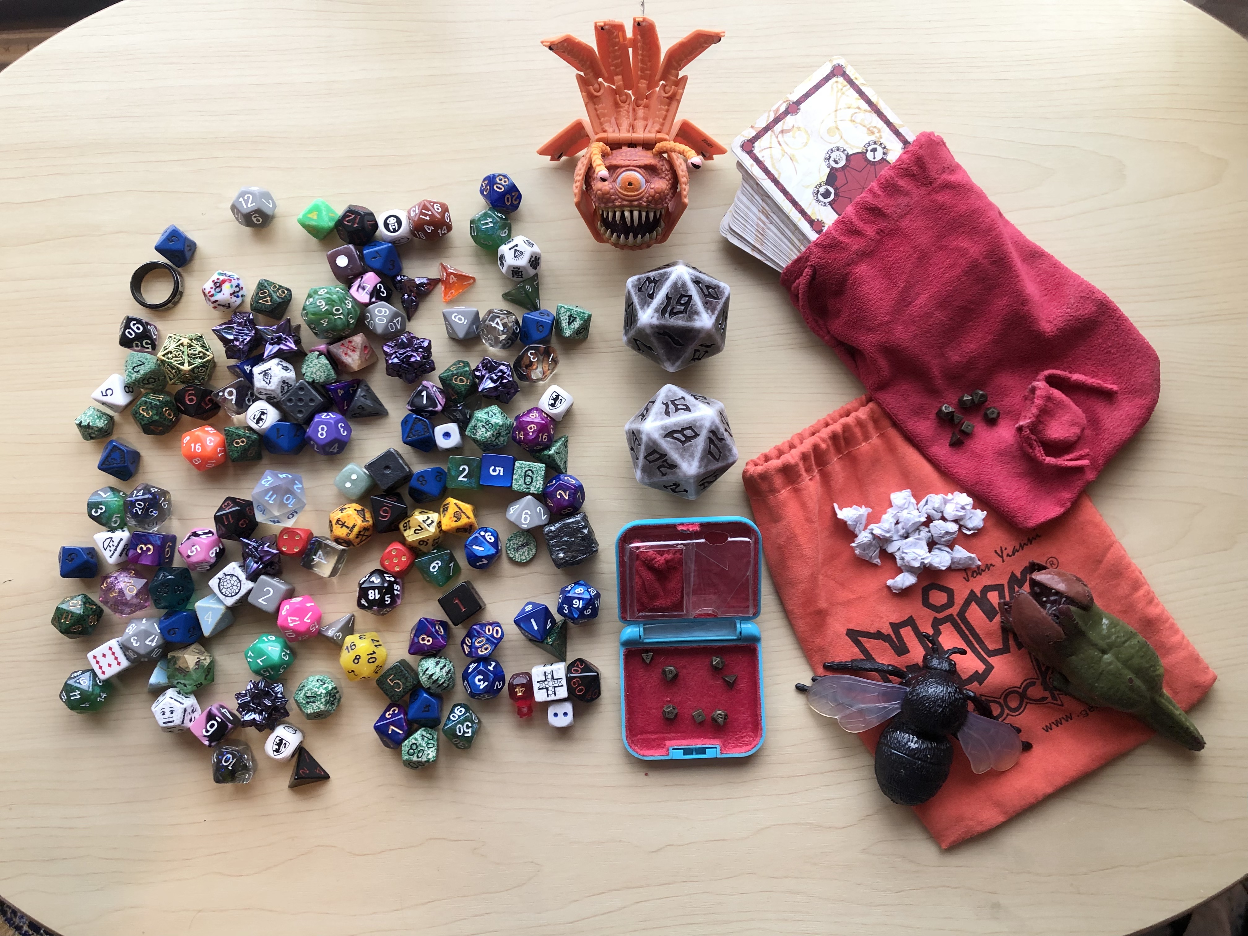 The dice collection, showing two giant grey chonk dice, the orange diceling of the beholder, the and the majority of the dice spread out inside the box lid of the d&d starter set. Beside it is the tiny dice roller box with tiny dice inside of it, the Hive tile game bag that acts as our dice bag, Simon and Lotus, the giant housefly and demodog, eating from a pile of scrap paper pieces, and the Harrow deck of many things, with tiny dice and dice bag on top of it.