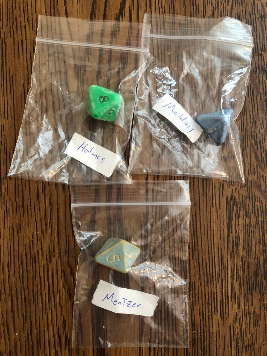 The original Holmes, Moldvay, and Mentzer dice inside separate ziplock bags, each one labelled. The Holmes is a heavily worn green d8 with black lettering, the Moldvay is a light blue d4 with white lettering, and the Mentzer is a slightly lighter blue d8 with white lettering, all sitting on a wooden table surface.