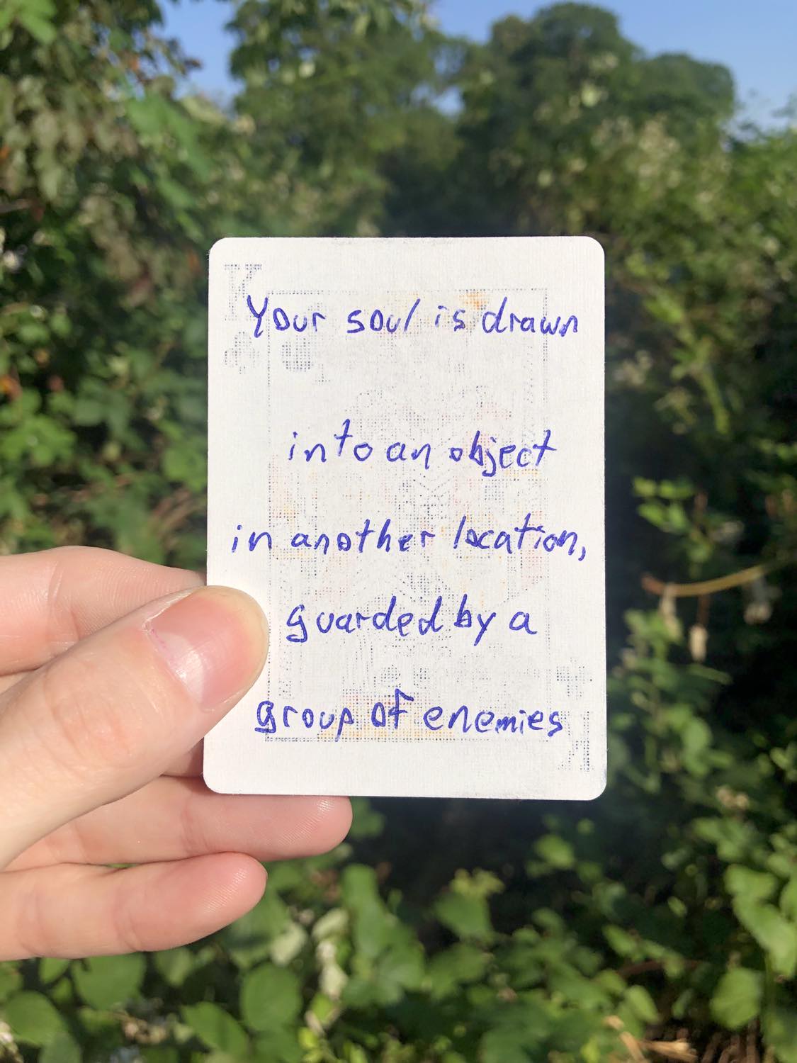 The king of clubs, held with the trees and foliage behind. Your soul is drawn into an object in another location, guarded by a group of enemies.