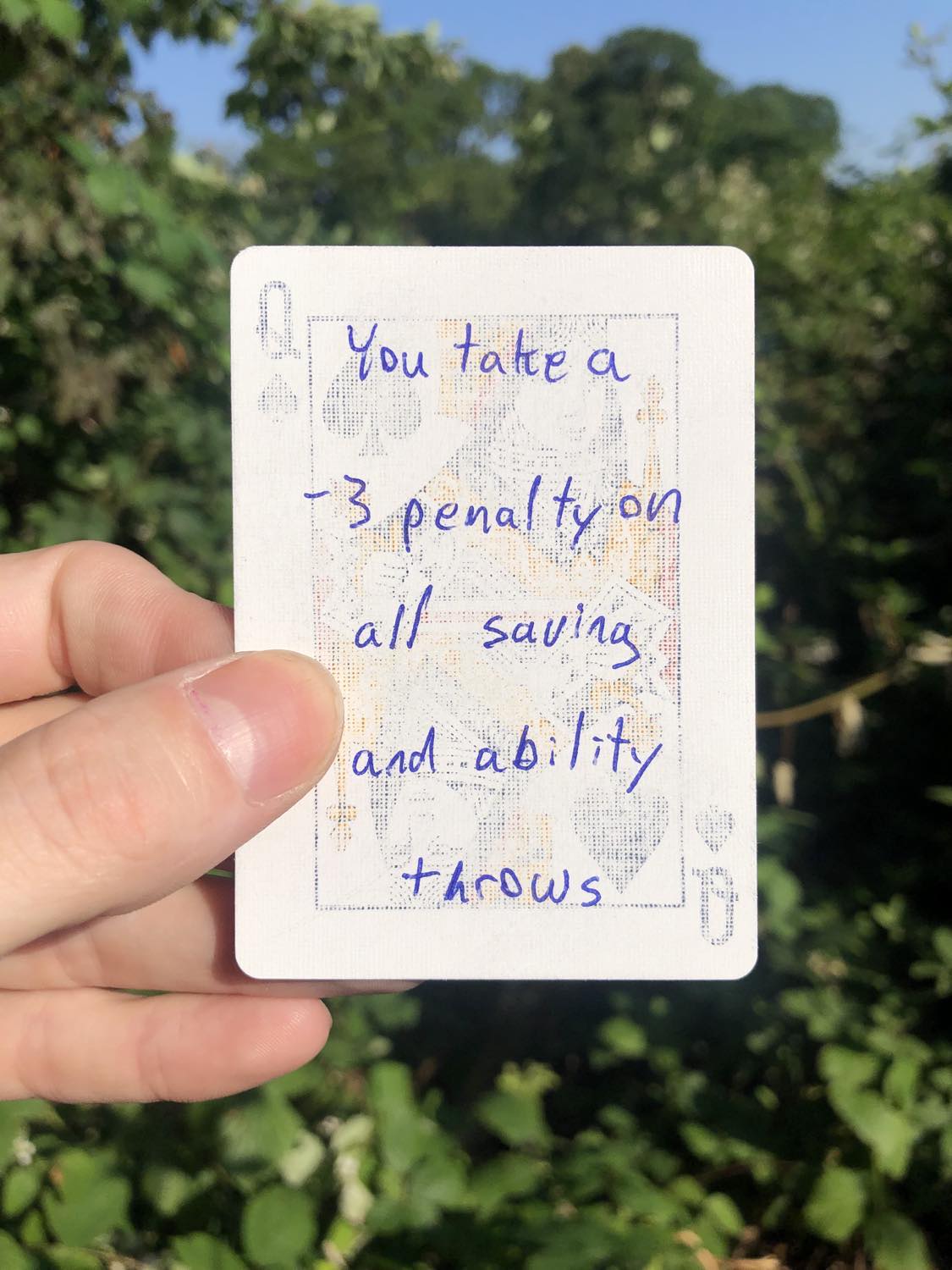 The queen of spades, held with the trees and foliage behind. You take a -3 penalty to all saving and ability throws.
