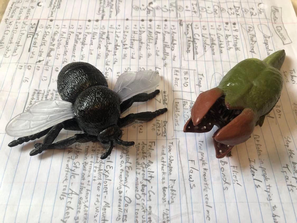 Simon and Lotus, a giant black housefly toy and a green and red demodog, or baby demogorgon toy, sitting on top of a character sheet for Mountain Dance, a tabaxi swarm ranger.